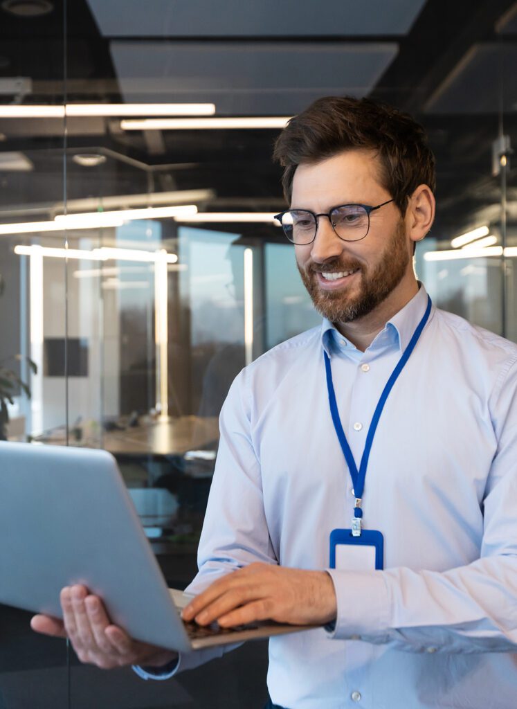 A male ShaodwHQ IT employee wearing their security badge while smiling down at the laptop they are holding open in their hands.