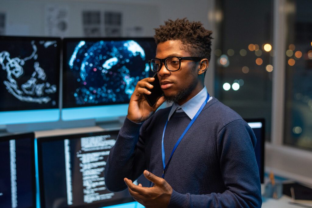 Young serious software developer or it-engineer consulting client on mobile phone while standing against his workplace.